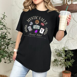 May include: Black t-shirt featuring a graphic print of the words "Mystic Falls Social Club" and various icons, including a vampire fang, a coffin, and a sign that says "Welcome to Mystic Falls, Virginia".
