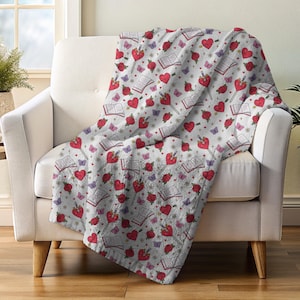 May include: A white throw blanket draped over a chair, featuring a repeating pattern of red hearts, roses, butterflies, and open books. The blanket has a soft, plush texture and is designed for comfort and warmth. The overall theme is romantic and whimsical.