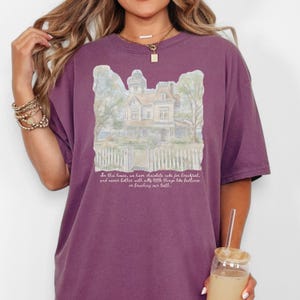 May include: A plum-colored short-sleeve t-shirt featuring a watercolor-style illustration of a house with a picket fence. Below the house, the text reads: In this house, we have chocolate cake for breakfast, and never bother with silly little things like bedtimes or brushing our teeth.