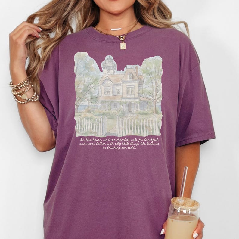 May include: A plum-colored short-sleeve t-shirt featuring a watercolor-style illustration of a house with a picket fence. Below the house, the text reads: In this house, we have chocolate cake for breakfast, and never bother with silly little things like bedtimes or brushing our teeth.