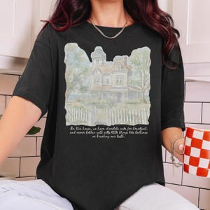 May include: A black t-shirt featuring a watercolor-style illustration of a large house with a white picket fence. Below the house, the text reads: In this house, we have chocolate cake for breakfast, and never bother with silly little things like bedtimes or brushing our teeth.
