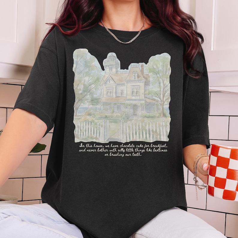 May include: A black t-shirt featuring a watercolor-style illustration of a large house with a white picket fence. Below the house, the text reads: In this house, we have chocolate cake for breakfast, and never bother with silly little things like bedtimes or brushing our teeth.