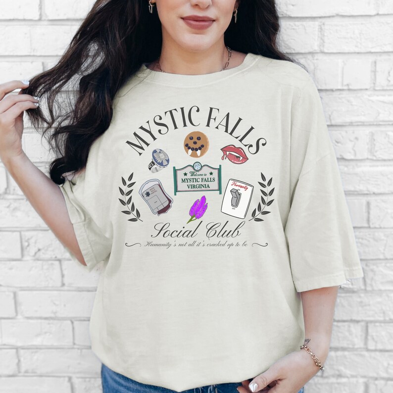 May include: A white t-shirt with a graphic design featuring the text "Mystic Falls Social Club" and various symbols, including a vampire fang, a blood bag, a flower, and a sign that reads "Welcome to Mystic Falls Virginia".