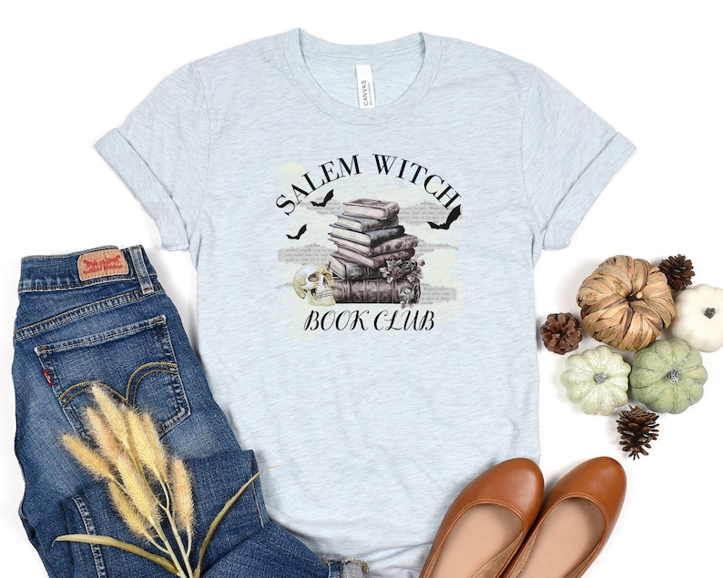 May include: Light blue t-shirt with the text "Salem Witch Book Club" in black lettering. A stack of books with a skull and bats are pictured above the text.