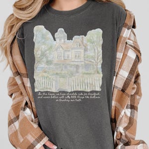 May include: A charcoal gray t-shirt featuring a watercolor illustration of a large house with a white picket fence. The text reads: In this house, we have chocolate cake for breakfast, and never bother with silly little things like bedtimes or brushing our teeth.