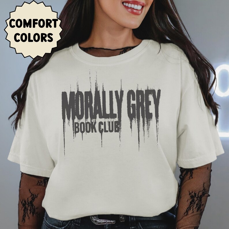 May include: Cream-colored t-shirt with the text "MORALLY GREY BOOK CLUB" in dark gray. The shirt has a crew neck and short sleeves. The words "COMFORT COLORS" are in a white circle in the upper left corner.