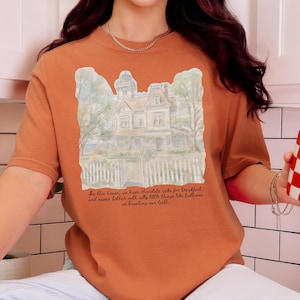 May include: A burnt orange t-shirt featuring a watercolor-style illustration of a Victorian house behind a white picket fence. Below the house, the text reads: In this house, we have chocolate cake for breakfast, and never bother with silly little things like bedtimes or brushing our teeth.