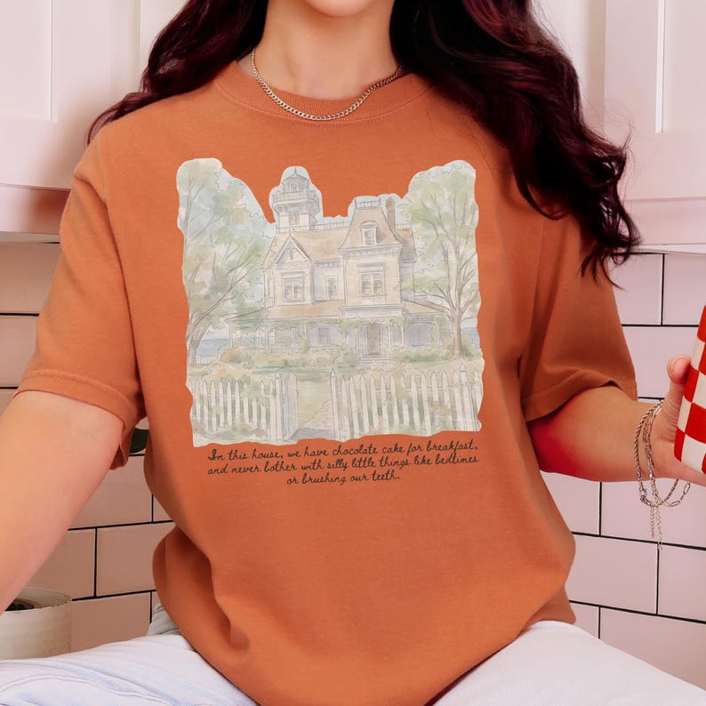 May include: A burnt orange t-shirt featuring a watercolor-style illustration of a Victorian house behind a white picket fence. Below the house, the text reads: In this house, we have chocolate cake for breakfast, and never bother with silly little things like bedtimes or brushing our teeth.