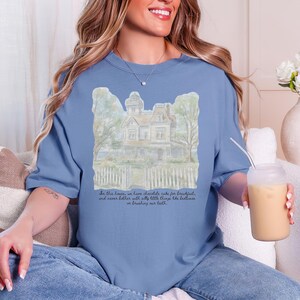 May include: A dusty blue crewneck t-shirt featuring a watercolor-style illustration of a Victorian house with a white picket fence. Below the house, the text reads: In this house, we have chocolate cake for breakfast, and never bother with silly little things like bedtimes or brushing our teeth.