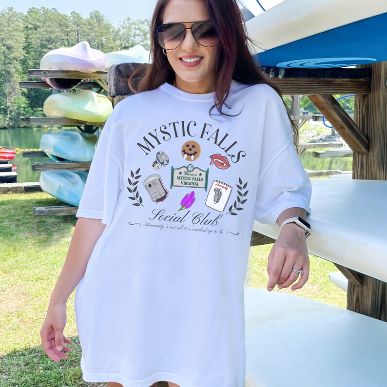 May include: White oversized t-shirt featuring a graphic print of the words "Mystic Falls Social Club" and various icons related to the TV show "The Vampire Diaries".