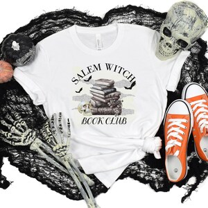 May include: White t-shirt with a black and white graphic design that says "Salem Witch Book Club". The graphic features a stack of books, bats, and a skull. The t-shirt is on a black and white background with a skull, pumpkins, and skeleton hands.