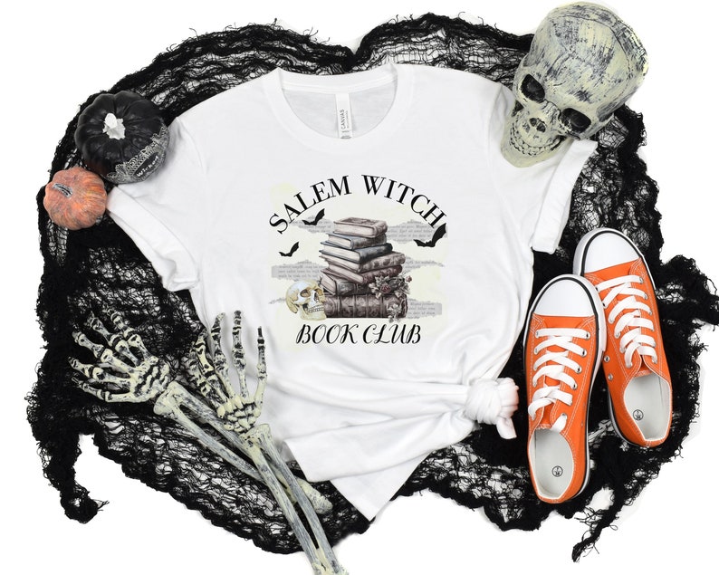 May include: White t-shirt with a black and white graphic design that says "Salem Witch Book Club". The graphic features a stack of books, bats, and a skull. The t-shirt is on a black and white background with a skull, pumpkins, and skeleton hands.