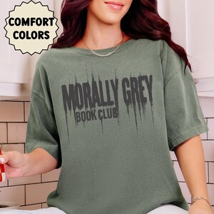 May include: Sage green t-shirt with the text "MORALLY GREY BOOK CLUB" in distressed black lettering. The shirt has short sleeves and a crew neck. The words "COMFORT COLORS" are in a circular design in the upper left corner.