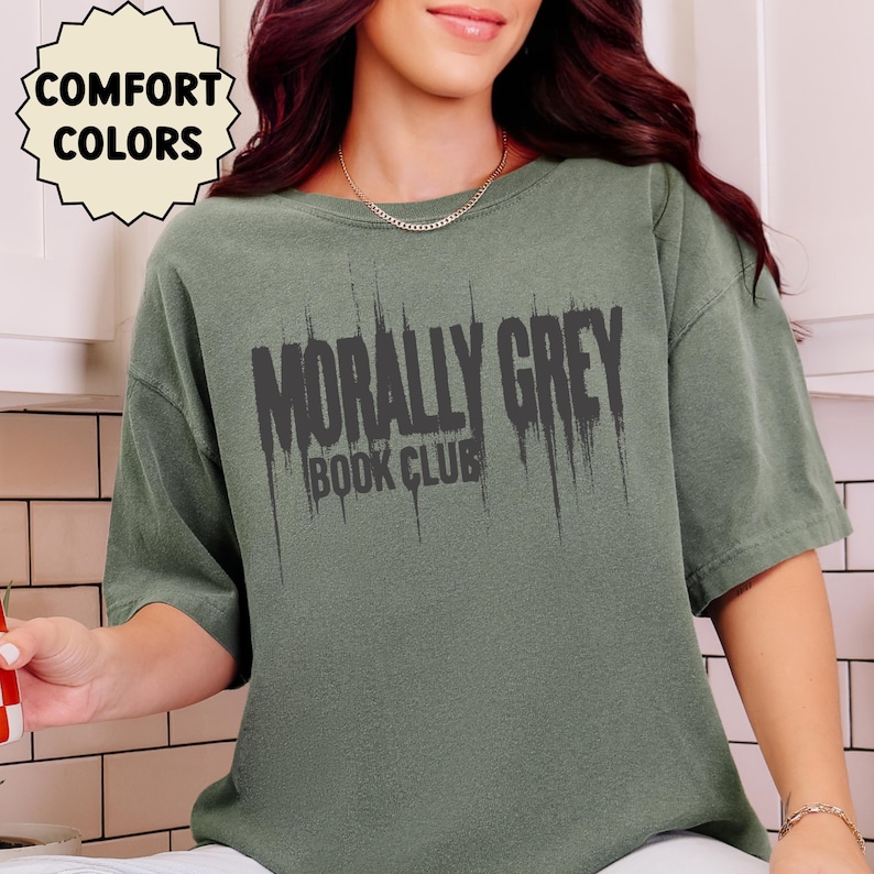 May include: Sage green t-shirt with the text "MORALLY GREY BOOK CLUB" in distressed black lettering. The shirt has short sleeves and a crew neck. The words "COMFORT COLORS" are in a circular design in the upper left corner.