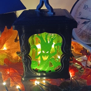 May include: A black lantern with a handle and a glass panel featuring a green silhouette of a spooky tree with a scary face. The lantern is illuminated from within, casting a warm glow.