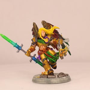 May include: A miniature figurine of a warrior with golden armor, blonde hair, and feathered wings. The warrior holds two swords, one green and one blue. The figure is posed on a circular base, with a detailed paint job.