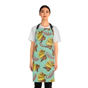 Peut inclure: Un tablier turquoise avec un motif répété de pintxos Gilda, avec des olives, des poivrons et des anchois. Le mot "Gilda" est écrit en cursive. Le tablier a une sangle de cou et des liens à la taille noirs.