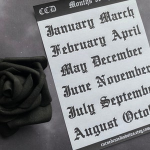 May include: A sheet of white paper with black gothic font stickers of the months of the year. The stickers are arranged in two rows. The text on the stickers reads: January, March, February, April, May, December, June, November, July, September, August, October. The text at the bottom of the sheet reads: cursedcutediabolus.etsy.com