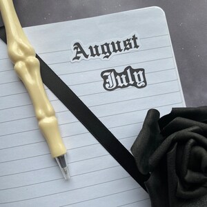 May include: A white notebook with black lines and a black ribbon bookmark. The notebook has two stickers on it, one that says "August" and one that says "July". A white skeleton pen is lying on the notebook. A black rose is in the bottom right corner of the image.