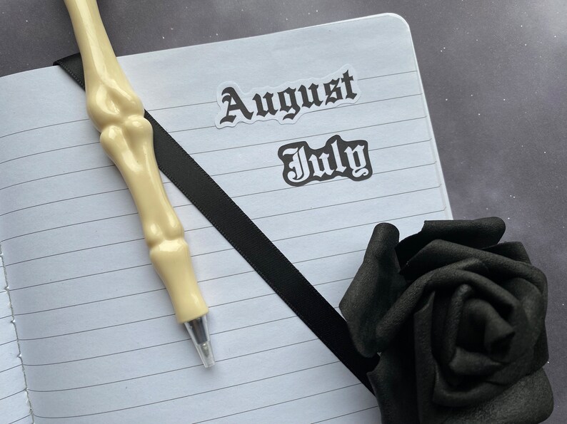 May include: A white notebook with black lines and a black ribbon bookmark. The notebook has two stickers on it, one that says "August" and one that says "July". A white skeleton pen is lying on the notebook. A black rose is in the bottom right corner of the image.