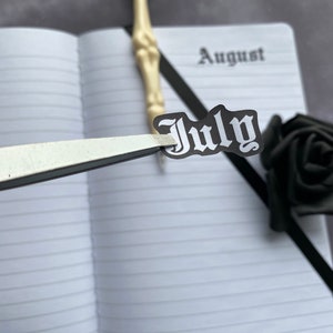 May include: A white notebook with black lined paper. The notebook has the word "August" written on the top of the page. A black and white sticker with the word "July" in a gothic font is being placed on the page with tweezers.