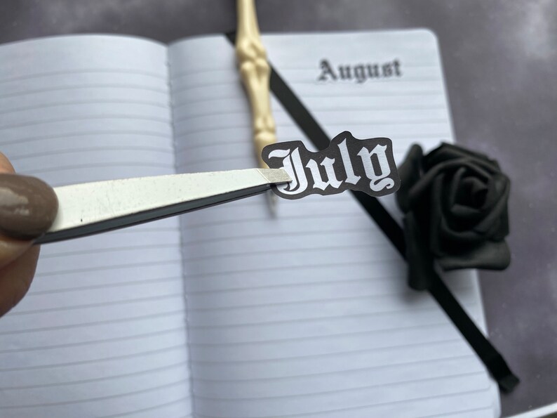 May include: A white notebook with black lined paper. The notebook has the word "August" written on the top of the page. A black and white sticker with the word "July" in a gothic font is being placed on the page with tweezers.