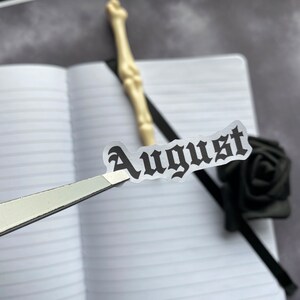 May include: A close-up of a sticker with the word "August" written in a gothic font. The sticker is being held by a pair of tweezers over a notebook.