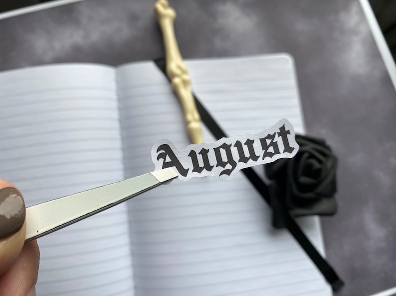 May include: A close-up of a sticker with the word "August" written in a gothic font. The sticker is being held by a pair of tweezers over a notebook.