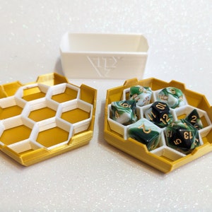 May include: A set of two hexagonal dice trays, one gold and one white. The gold tray has a honeycomb design and is filled with green and white dice with gold numbers. The white tray is empty.