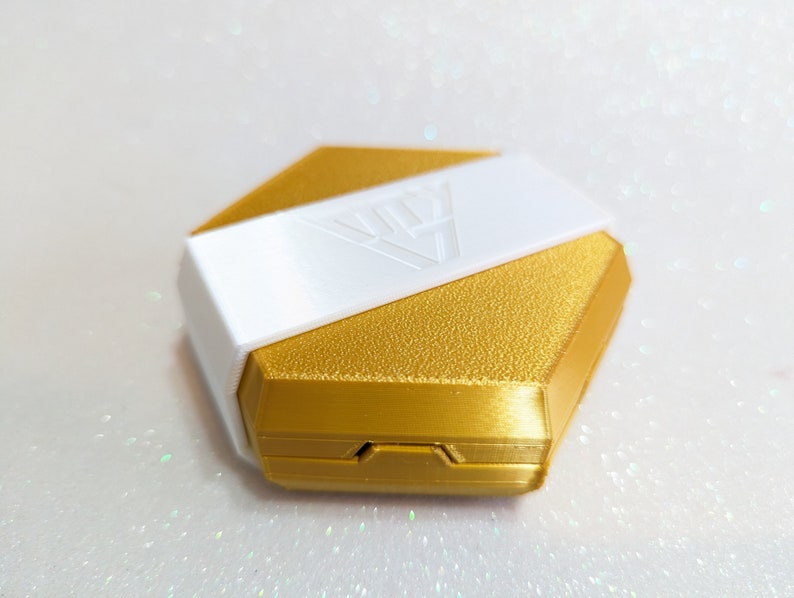 May include: A gold hexagonal box with a white lid. The lid has a geometric design and the box has a textured surface.