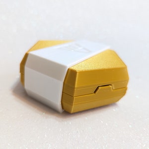 May include: A hexagonal shaped box with a gold top and a white bottom. The box is closed and has a small hinge on the side.