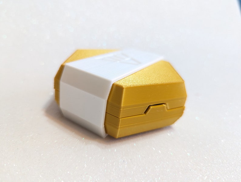May include: A hexagonal shaped box with a gold top and a white bottom. The box is closed and has a small hinge on the side.