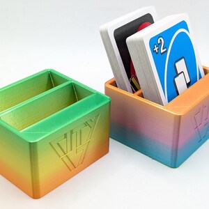 May include: Two 3D printed card holders with a gradient colour scheme. The holder on the left has two compartments and the holder on the right has one compartment. The holder on the right has a deck of UNO cards in it. The holder on the right has the text "DIY" on the front.