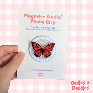 May include: A magnetic Kindle and phone grip featuring a monarch butterfly design with orange and black wings. The product is mounted on a white display card with the text Magnetic Kindle/Phone Grip, MagSafe Compatible, and Oodles and Doodles.