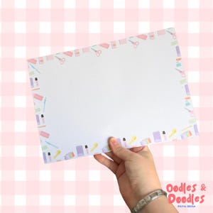 May include: A rectangular sheet of white paper featuring a dotted grid pattern and a decorative border composed of small, colourful illustrations of stationery items including scissors, pens, erasers, and notebooks in pastel shades of pink, blue, and purple.