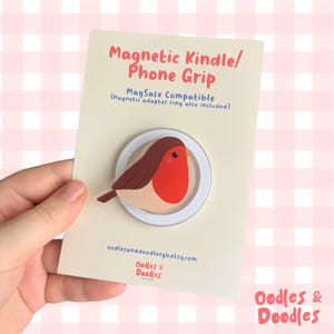 May include: A magnetic Kindle and phone grip on a cream-colored display card. The grip features a robin illustration with a red chest and brown wings. Text on the card reads: Magnetic Kindle/Phone Grip, MagSafe Compatible, and oodlesanddoodlesgb.etsy.com.