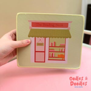 May include: A rectangular mousepad with a bookstore illustration. The design shows a red and pink shop front with the text "The Reading Nook" above the awning. Inside, there are bookshelves and an orange cat. The mousepad has a light yellow border.