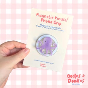 May include: A purple octopus phone grip on a white card with the text "Magnetic Kindle/Phone Grip". The grip is MagSafe compatible and includes a magnetic adapter ring. The card has the shop name "Oodles & Doodles" and a pink and white checkered background.