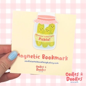 A hand holding a magnetic bookmark of a pink and green jar of pickles with a label saying "Whatever tickles your pickle!" on a square backing card.