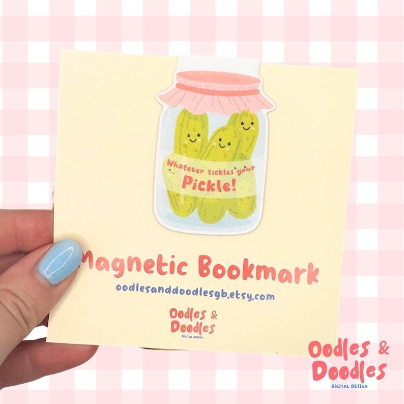 A hand holding a magnetic bookmark of a pink and green jar of pickles with a label saying "Whatever tickles your pickle!" on a square backing card.