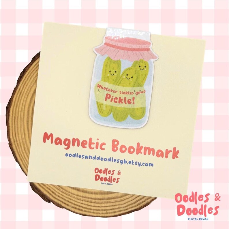 A hand holding a magnetic bookmark of a pink and green jar of pickles with a label saying "Whatever tickles your pickle!" on a square backing card.
