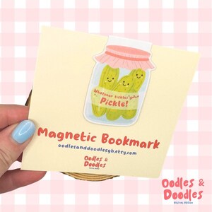 A hand holding a magnetic bookmark of a pink and green jar of pickles with a label saying "Whatever tickles your pickle!" on a square backing card.