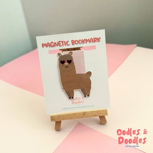 May include: A brown llama with pink heart-shaped sunglasses on a white magnetic bookmark. The bookmark has the text "Magnetic Bookmark" at the top and "Oodles & Doodles" at the bottom. The bookmark is on a small wooden easel.