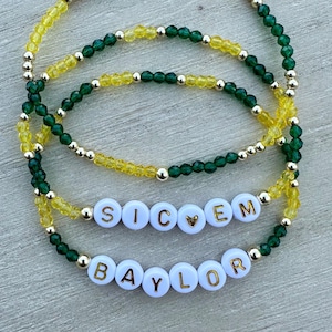 May include: Three layered beaded bracelets with green and yellow beads. The bracelets spell out "SIC 'EM BAYLOR" in white letters with gold accents.