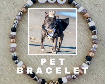 May include: A gold charm bracelet with black, white, and brown beads. The charm is a gold paw print. The bracelet has white beads with the letters "G" and two hearts. The bracelet is displayed on a wooden surface with a photo of a dog wearing a tuxedo and a crown.