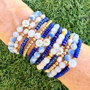 May include: A stack of ten beaded bracelets featuring a mix of blue and white marbled beads, small gold-toned metallic spheres, and white circular beads with gold-colored letter V or cross symbols printed on the surface in a repeating pattern.