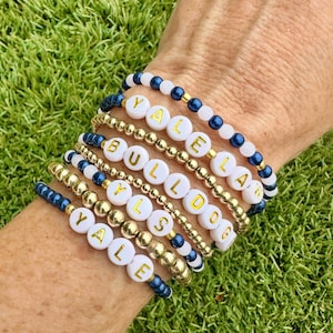 May include: Several beaded bracelets spelling "YALE" and "BULLDOGS" in gold lettering on white beads. The bracelets are a mix of gold, navy blue, and white beads. The bracelets are stacked on a wrist against a background of green grass.