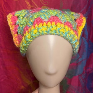 May include: A colorful, handmade crochet hat in a granny square pattern. The hat features vibrant pink, yellow, and turquoise yarn. The hat is displayed on a mannequin head. This item is perfect for adding a pop of color to any outfit.