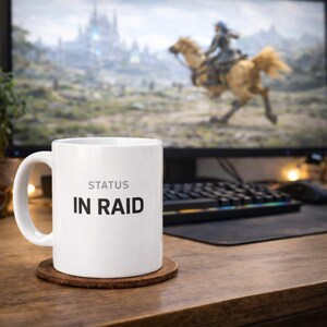 Puede incluir: Taza de cerámica blanca con el texto "STATUS IN RAID" en negro, sobre un posavasos marrón. Un monitor de ordenador que muestra una escena de videojuego está en el fondo, junto con un teclado y un ratón.