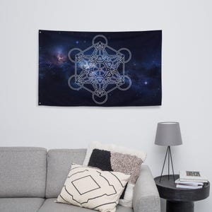Op de afbeelding: Een rechthoekige wandtapijt met een wit Metatron's Cube-ontwerp tegen een donkerblauwe en paarse melkwegachtergrond. Het tapijt hangt boven een grijze bank met decoratieve kussens.